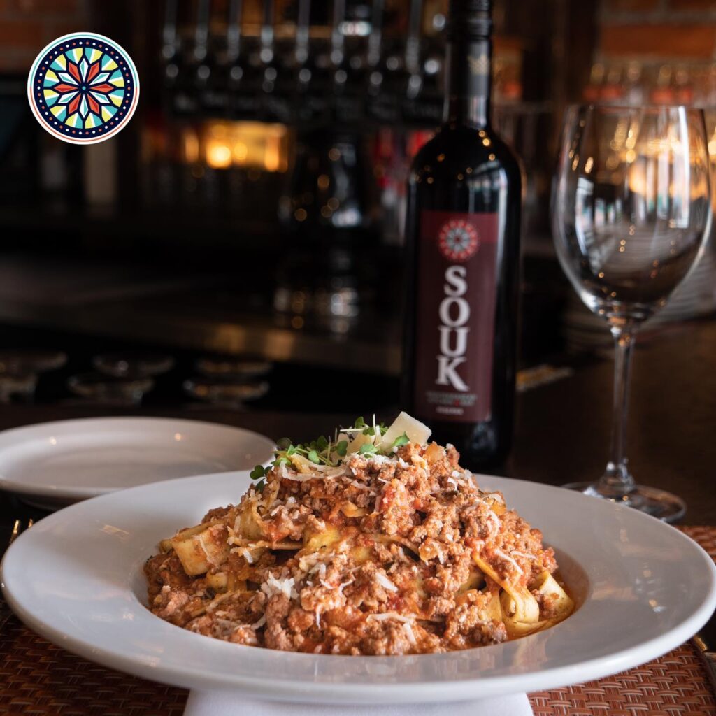 A photo of a delicious looking Bolognese next to a bottle of wine in a low lit dining room.