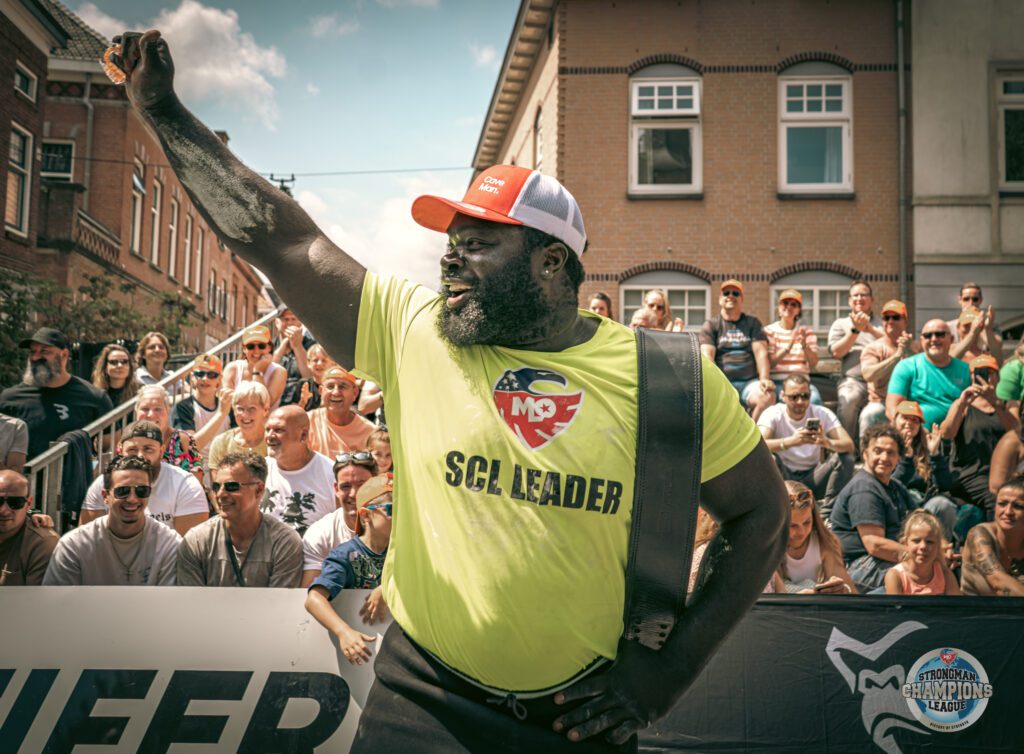 Strongman raises his right fist in the air in celebration as a crowd smiles in the background